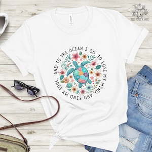 Puede incluir: Camiseta blanca con un diseño de tortuga marina colorida rodeada de flores y el texto "And to the ocean I go, to lose my mind, and find my soul." La camiseta también tiene el texto "Thunder & Grace Tees, Totes, & More."