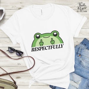 May include: White t-shirt featuring a green frog graphic with the text "RESPECTFULLY". The frog is giving the middle finger with both hands. The shirt is a crew neck and has short sleeves. The brand name "Thunder & Grace" is printed on the upper right.
