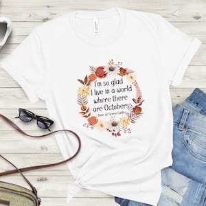May include: White t-shirt with a floral wreath design and the quote "I'm so glad I live in a world where there are Octobers" from Anne of Green Gables. The shirt is paired with blue jeans and accessories.
