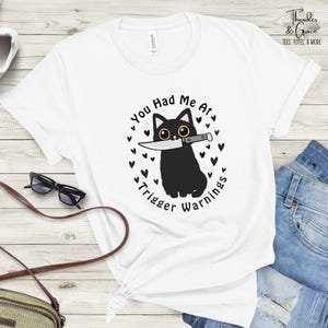 May include: White t-shirt featuring a black cat holding a knife with the text "You Had Me At Trigger Warnings." The cat is surrounded by heart shapes. The shirt is a casual wear item.