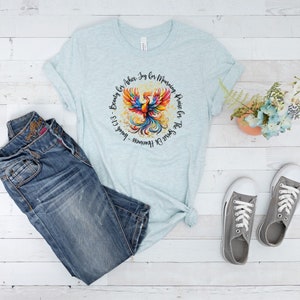 May include: A light blue t-shirt with a colorful phoenix design and the text "Spirit Of Heaviness - Isaiah 61:3 - Beauty For Ashes. Joy For Mourning. Praise For The Soul Of The"