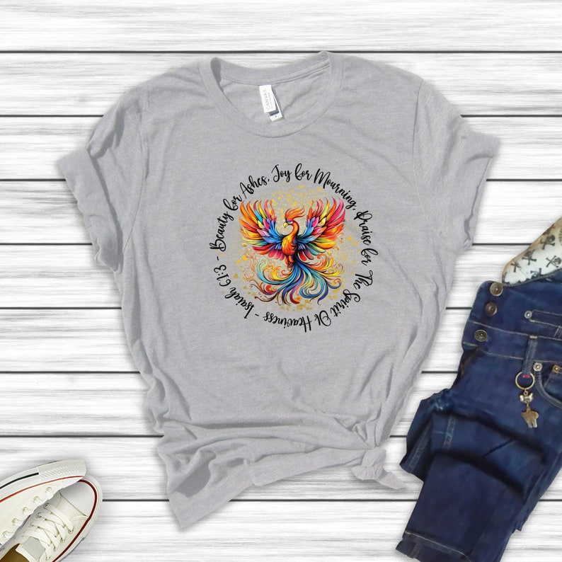 May include: A gray t-shirt with a colorful phoenix design and the text "The Spirit of Heaviness -  Beauty for Ashes. Joy for Mourning. Praise for The Spirit of Heaviness - Isaiah 61:3"