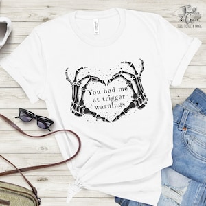 May include: White t-shirt with a black graphic of skeleton hands forming a heart shape, with the text "You had me at trigger warnings." The shirt also has the text "Thunder & Grace Tees, Totes, & More."