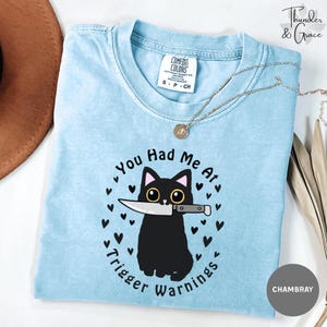 May include: A light blue short-sleeve t-shirt with a graphic of a black cat holding a knife in its mouth. The text reads You Had Me At Trigger Warnings, surrounded by small black hearts. The shirt features a Comfort Colors brand neck label.
