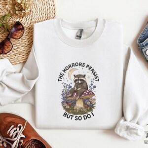 Puede incluir: Sudadera blanca con un diseño de un mapache sentado en una seta, rodeado de flores y una luna creciente. El texto dice "THE HORRORS PERSIST BUT SO DO I." La sudadera es de cuello redondo.
