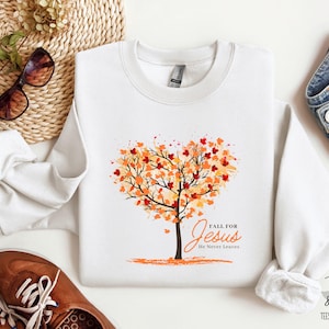 Könnte beinhalten: Weißer Pullover mit einem herbstlichen Design, das einen Baum mit orangefarbenen und roten Blättern in Form eines Herzens zeigt. Der Text "FALL FOR Jesus He Never Leaves" ist unter dem Baum gedruckt.