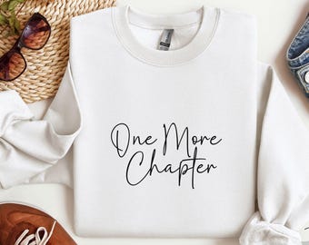 May include: A white crew neck sweatshirt featuring the text "One More Chapter" in black script. The image includes sunglasses, a woven basket, and brown shoes. The sweatshirt is a casual style.