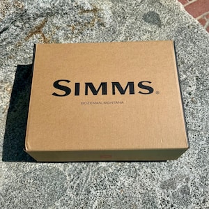 May include: A brown cardboard box with the brand name "SIMMS" printed in black, along with "BOZEMAN, MONTANA" below it. The box is rectangular and sits on a gray stone surface.