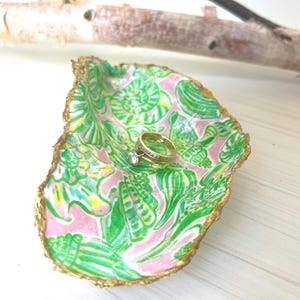May include: A pink and green patterned ceramic ring dish with a gold rim. The dish is shaped like a seashell and has a ring resting inside.