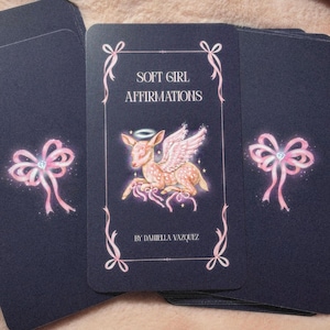 May include: A deck of navy blue affirmation cards with pink bow and diamond accents. The center card features a winged deer illustration with the text "SOFT GIRL AFFIRMATIONS" and "BY DANIELLA VAZQUEZ".