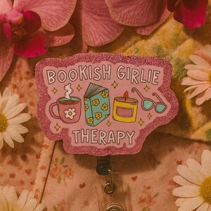 Puede incluir: Un portatarjetas rosa brillante con el texto "BOOKISH GIRLIE THERAPY" e ilustraciones de una taza de caf&eacute;, un libro, una vela y gafas. El portatarjetas est&aacute; rodeado de flores rosas y margaritas.