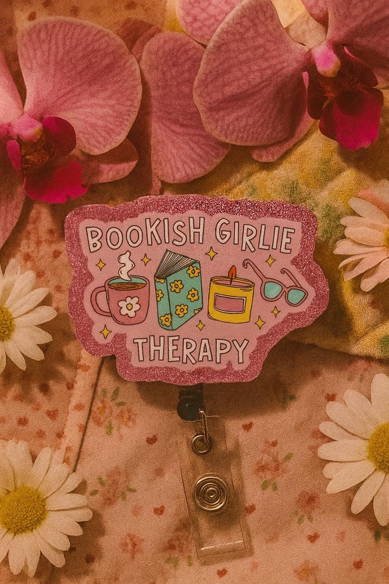 Puede incluir: Un portatarjetas rosa brillante con el texto "BOOKISH GIRLIE THERAPY" e ilustraciones de una taza de caf&eacute;, un libro, una vela y gafas. El portatarjetas est&aacute; rodeado de flores rosas y margaritas.