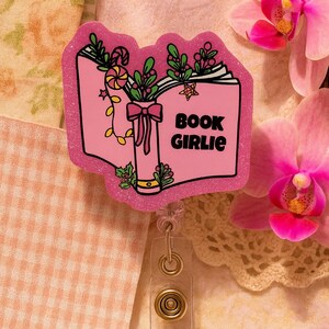 Puede incluir: Portatarjetas rosa brillante con dise&ntilde;o de libro. El libro tiene las palabras "BOOK GIRLIE" en la portada, con elementos decorativos de hojas, caramelos y un lazo. El portatarjetas tiene un clip de pl&aacute;stico transparente y un cierre met&aacute;lico.
