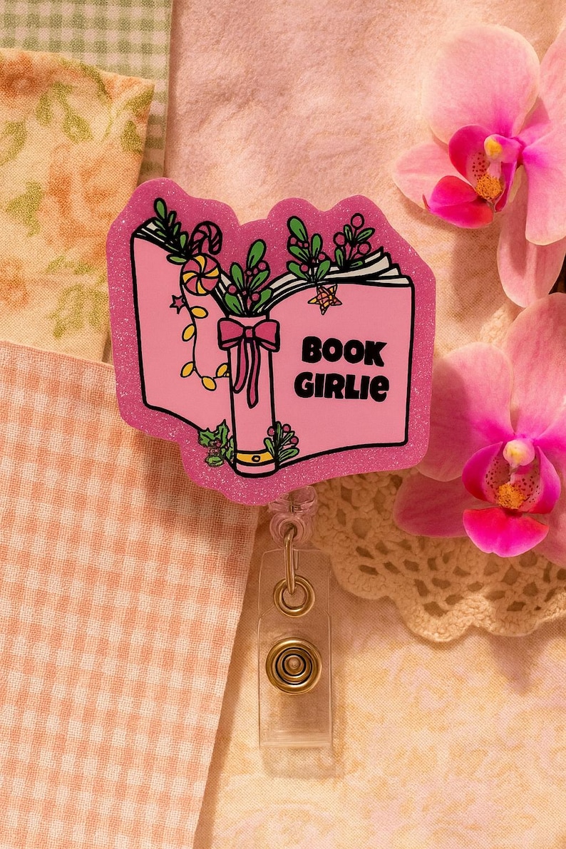 Puede incluir: Portatarjetas rosa brillante con dise&ntilde;o de libro. El libro tiene las palabras "BOOK GIRLIE" en la portada, con elementos decorativos de hojas, caramelos y un lazo. El portatarjetas tiene un clip de pl&aacute;stico transparente y un cierre met&aacute;lico.