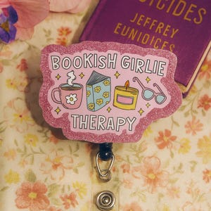 Puede incluir: Un portatarjetas rosa brillante con el texto "BOOKISH GIRLIE THERAPY" e ilustraciones de una taza de caf&eacute;, un libro, una vela y gafas. Un libro morado titulado "THE VIRGIN SUICIDES" est&aacute; en el fondo.