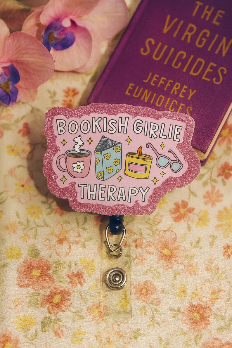 Puede incluir: Un portatarjetas rosa brillante con el texto "BOOKISH GIRLIE THERAPY" e ilustraciones de una taza de caf&eacute;, un libro, una vela y gafas. Un libro morado titulado "THE VIRGIN SUICIDES" est&aacute; en el fondo.