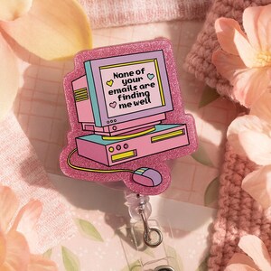 Puede incluir: Un portatarjetas en forma de ordenador rosa y morado con el texto "None of your emails are finding me well". El portatarjetas tiene un cord&oacute;n retr&aacute;ctil y un clip de pl&aacute;stico transparente. El dise&ntilde;o incluye un monitor de ordenador, una base de ordenador y un rat&oacute;n.