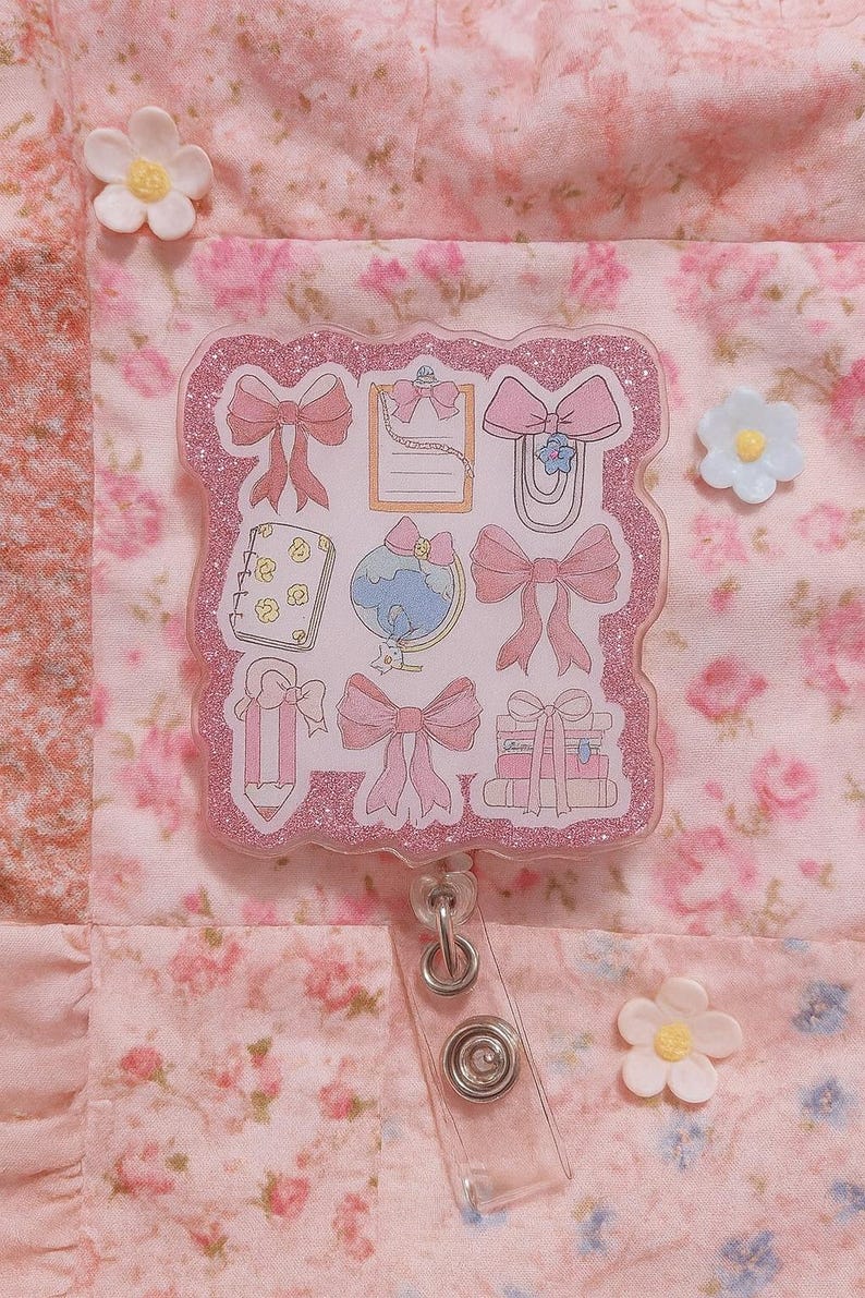 Puede incluir: Un portatarjetas rosa brillante con ilustraciones de lazos rosas, un globo terr&aacute;queo y un bloc de notas. Sobre un fondo floral rosa con detalles de flores blancas.