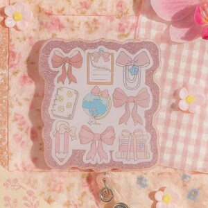 Puede incluir: Un portatarjetas rosa brillante con ilustraciones de lazos rosas y art&iacute;culos de papeler&iacute;a. El portatarjetas tiene un cord&oacute;n retr&aacute;ctil de pl&aacute;stico transparente y un clip met&aacute;lico. El fondo es un estampado floral y de cuadros vichy.