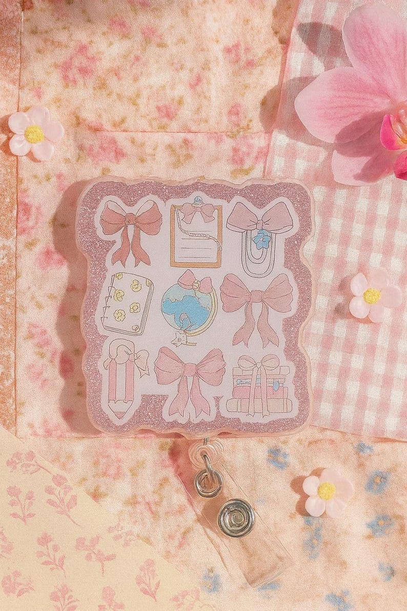Puede incluir: Un portatarjetas rosa brillante con ilustraciones de lazos rosas y art&iacute;culos de papeler&iacute;a. El portatarjetas tiene un cord&oacute;n retr&aacute;ctil de pl&aacute;stico transparente y un clip met&aacute;lico. El fondo es un estampado floral y de cuadros vichy.