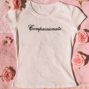 Könnte beinhalten: Ein weißes T-Shirt mit dem Wort "Compassionate" in kursiver Schrift. Das Shirt ist von rosa Rosen und kleinen rosa Blumen auf einem rosa Hintergrund mit einem Karomuster umgeben.