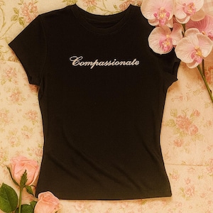 May include: Black t-shirt with the word "Compassionate" embroidered in white script. The shirt is displayed on a floral background with pink roses and orchids. A simple, elegant design.