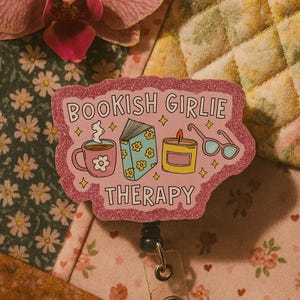 Puede incluir: Una insignia rosa brillante con las palabras "BOOKISH GIRLIE THERAPY" e ilustraciones de una taza, un libro, una vela y gafas. La insignia est&aacute; sobre un fondo floral con una flor rosa.