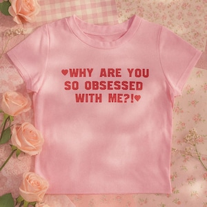 May include: A light pink t-shirt with the text "WHY ARE YOU SO OBSESSED WITH ME?!" in red. The shirt is surrounded by pink roses and floral patterned fabric.
