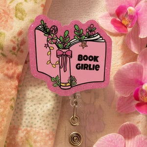 Puede incluir: Un portatarjetas rosa brillante con un dise&ntilde;o de libro abierto y las palabras "BOOK GIRLIE". El libro est&aacute; decorado con hojas verdes, un lazo y bastones de caramelo. El carrete tiene una correa de pl&aacute;stico transparente y un clip de metal.