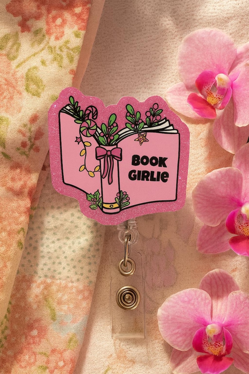 Puede incluir: Un portatarjetas rosa brillante con un dise&ntilde;o de libro abierto y las palabras "BOOK GIRLIE". El libro est&aacute; decorado con hojas verdes, un lazo y bastones de caramelo. El carrete tiene una correa de pl&aacute;stico transparente y un clip de metal.