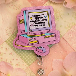 Puede incluir: Un portatarjetas en forma de ordenador rosa y morado con el texto "None of your emails are finding me well." El portatarjetas tiene un cord&oacute;n retr&aacute;ctil y un clip de pl&aacute;stico transparente. El fondo es un edred&oacute;n de retazos con flores rosas.