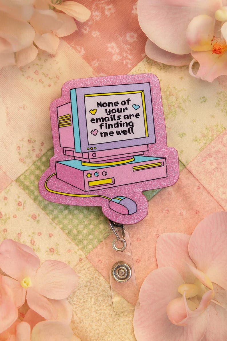 Puede incluir: Un portatarjetas en forma de ordenador rosa y morado con el texto "None of your emails are finding me well." El portatarjetas tiene un cord&oacute;n retr&aacute;ctil y un clip de pl&aacute;stico transparente. El fondo es un edred&oacute;n de retazos con flores rosas.