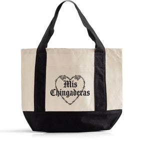K&ouml;nnte beinhalten: Cremefarbene Tragetasche mit schwarzen Griffen und Boden. Die Tasche zeigt eine schwarze Herzgrafik mit den Worten "Mis Chingaderas" in gotischer Schrift. Das Design ist mittig auf der Vorderseite.