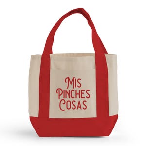 May include: A canvas tote bag with a red base, handles, and trim. The main body is a natural beige color, with a front pocket. The words "MIS PINCHES COSAS" are printed in red script on the front of the bag.