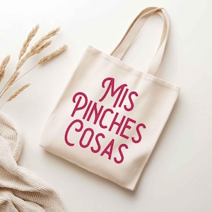 K&ouml;nnte beinhalten: Eine cremefarbene Tragetasche mit dem Text "Mis Pinches Cosas" in einer kr&auml;ftigen, kursiven, magenta Schrift. Die Tasche hat zwei Griffe und ist aus einem robusten Stoff gefertigt. Der Hintergrund ist wei&szlig;, mit einem Zweig aus getrocknetem Weizen und einer cremefarbenen Strickdecke.