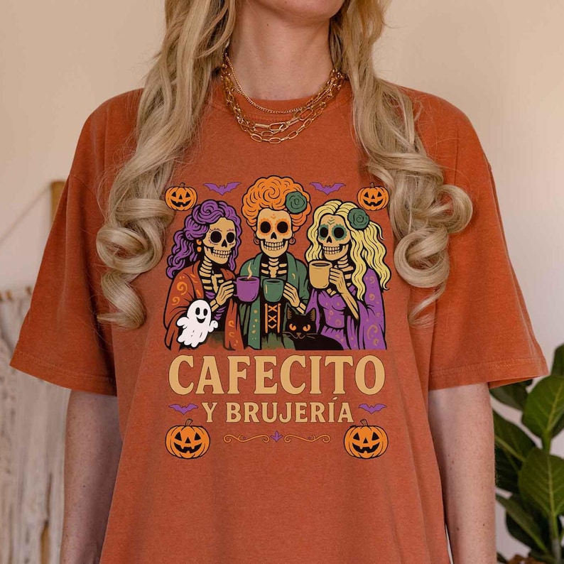 May include: Burnt orange t-shirt featuring a graphic of three skeleton witches holding mugs, a ghost, and a black cat. The text "CAFECITO Y BRUJER&Iacute;A" is printed below the graphic, with pumpkin and bat illustrations.