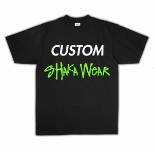 May include: A black short-sleeved t-shirt featuring the word CUSTOM printed in white capital letters on the upper chest, with the words SHAKA WEAR printed below in a bright lime green stylised font across the centre of the garment.