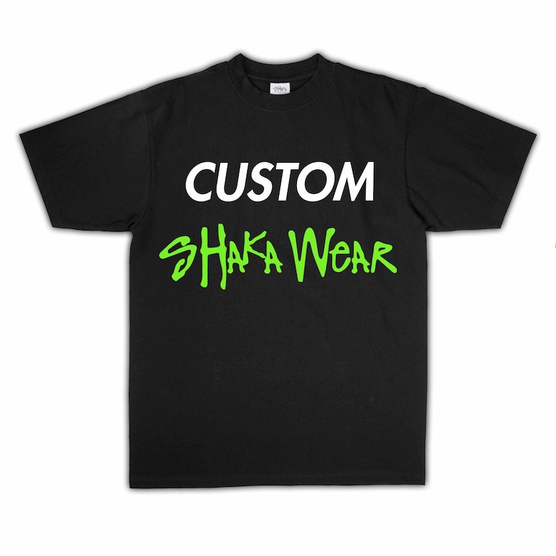 May include: A black short-sleeved t-shirt featuring the word CUSTOM printed in white capital letters on the upper chest, with the words SHAKA WEAR printed below in a bright lime green stylised font across the centre of the garment.