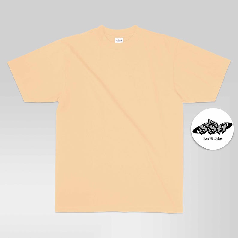 May include: A solid light peach-coloured short-sleeve t-shirt with a crew neckline. A small white circular graphic is positioned to the right, featuring the black text SSA and Los Angeles in a stylised font inside a black oval shape.