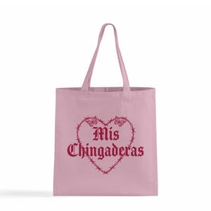 K&ouml;nnte beinhalten: Eine hellrosa Tragetasche mit passenden Griffen. Die Tasche zeigt ein herzf&ouml;rmiges Design aus Stacheldraht und Schmetterlingen mit den Worten "Mis Chingaderas" in burgunderfarbener Schrift.