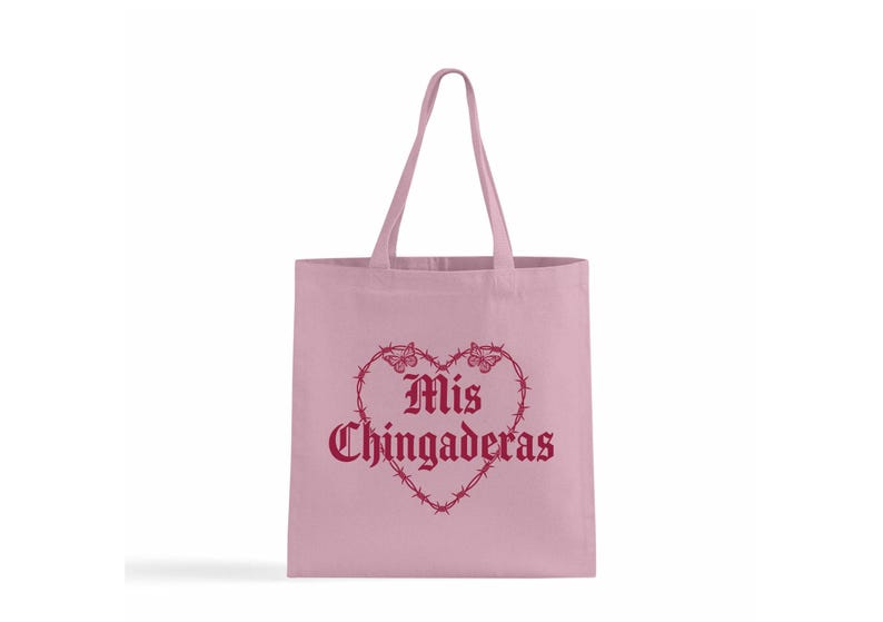 K&ouml;nnte beinhalten: Eine hellrosa Tragetasche mit passenden Griffen. Die Tasche zeigt ein herzf&ouml;rmiges Design aus Stacheldraht und Schmetterlingen mit den Worten "Mis Chingaderas" in burgunderfarbener Schrift.