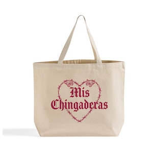 K&ouml;nnte beinhalten: Beige Canvas-Tragetasche mit herzf&ouml;rmigem Design aus Stacheldraht und Schmetterlingen. Die Worte "Mis Chingaderas" sind in Burgunderrot im Herzen aufgedruckt. Die Tasche hat zwei Griffe.