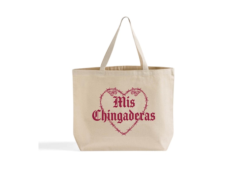 K&ouml;nnte beinhalten: Beige Canvas-Tragetasche mit herzf&ouml;rmigem Design aus Stacheldraht und Schmetterlingen. Die Worte "Mis Chingaderas" sind in Burgunderrot im Herzen aufgedruckt. Die Tasche hat zwei Griffe.