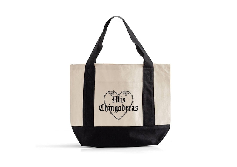 K&ouml;nnte beinhalten: Cremefarbene Tragetasche mit schwarzen Griffen und Boden. Die Tasche zeigt eine schwarze Herzgrafik mit den Worten "Mis Chingaderas" in gotischer Schrift. Das Design ist mittig auf der Vorderseite.