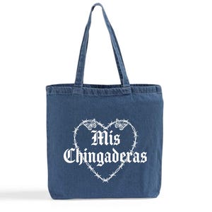 K&ouml;nnte beinhalten: Eine blaue Denim-Tragetasche mit einer wei&szlig;en herzf&ouml;rmigen Stacheldraht-Design mit Schmetterlingen und den Worten "Mis Chingaderas" in gotischer Schrift. Die Tasche hat zwei Griffe.