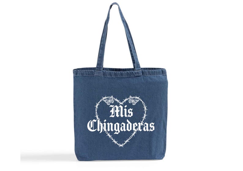 K&ouml;nnte beinhalten: Eine blaue Denim-Tragetasche mit einer wei&szlig;en herzf&ouml;rmigen Stacheldraht-Design mit Schmetterlingen und den Worten "Mis Chingaderas" in gotischer Schrift. Die Tasche hat zwei Griffe.