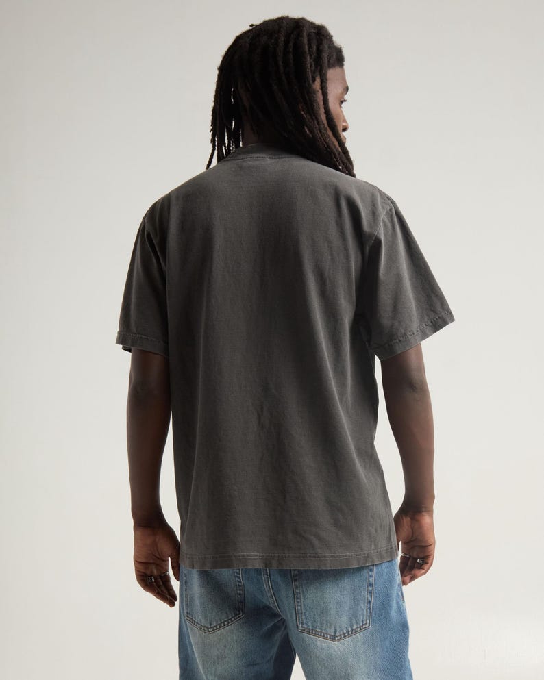 May include: A short-sleeved, charcoal grey t-shirt with a crew neckline, worn with blue denim jeans. The shirt features a solid, plain back design with a relaxed fit, showcasing a faded, washed-out fabric texture and standard hemline construction.