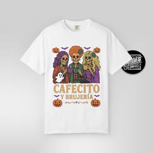 May include: White t-shirt featuring a Halloween-themed graphic. The design includes three skeleton figures with colorful hair, holding cups, and a ghost. The text "CAFECITO Y BRUJER&Iacute;A" is displayed below the figures, with pumpkins and bats.