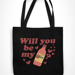 Op de afbeelding: Zwarte draagtas met de tekst "Will you be my" in het rood, een fles Valentina-chilisaus en hartjes. Een item met Valentijnsdag-thema.