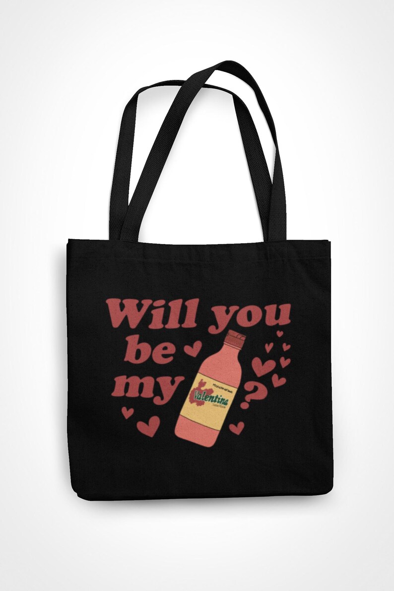 Op de afbeelding: Zwarte draagtas met de tekst "Will you be my" in het rood, een fles Valentina-chilisaus en hartjes. Een item met Valentijnsdag-thema.
